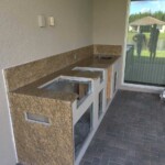 Outdoor kitchen countertop with built-in grill and sink under a covered patio.